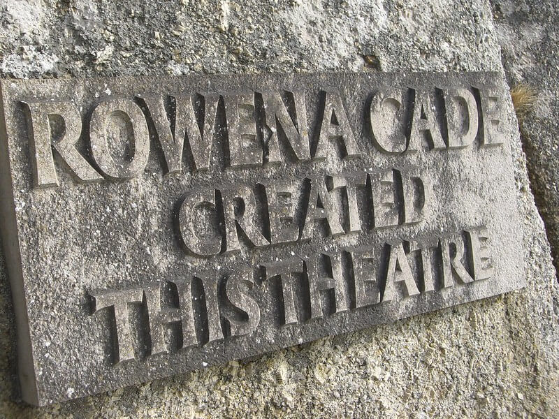 A stone plaque on a rough wall reads, ROWENA CADE CREATED THIS THEATRE, with deeply engraved, weathered lettering at the iconic Minack Theatre.