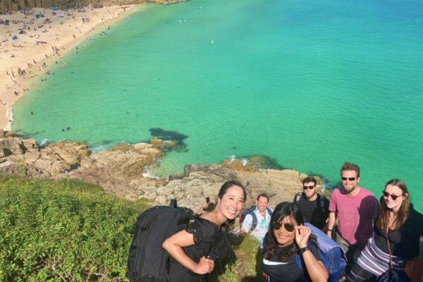 A group of people smile at the camera on a grassy cliff during a coast walk from Zennor to St Ives, overlooking a turquoise sea and sandy beach, with others relaxing and swimming below on a sunny day.