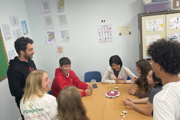 Seven teenagers sit and stand around a rectangular table playing a board game in the classroom, which is decorated with colorful student drawings and educational posters during their english language classes.
