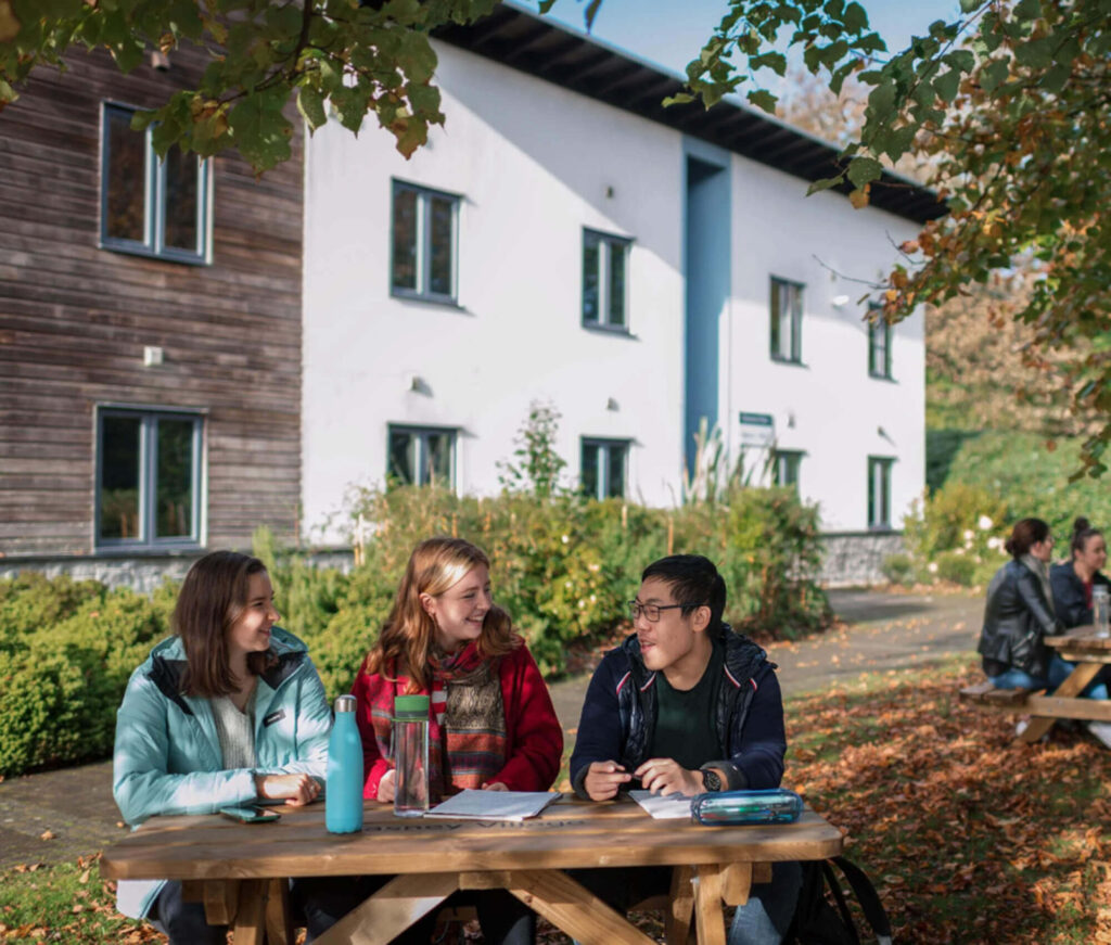 Three students sit at a wooden picnic table outside their school, smiling and talking, with books and notebooks in front of them. Colorful autumn trees frame the location, while a modern building rises in the background.