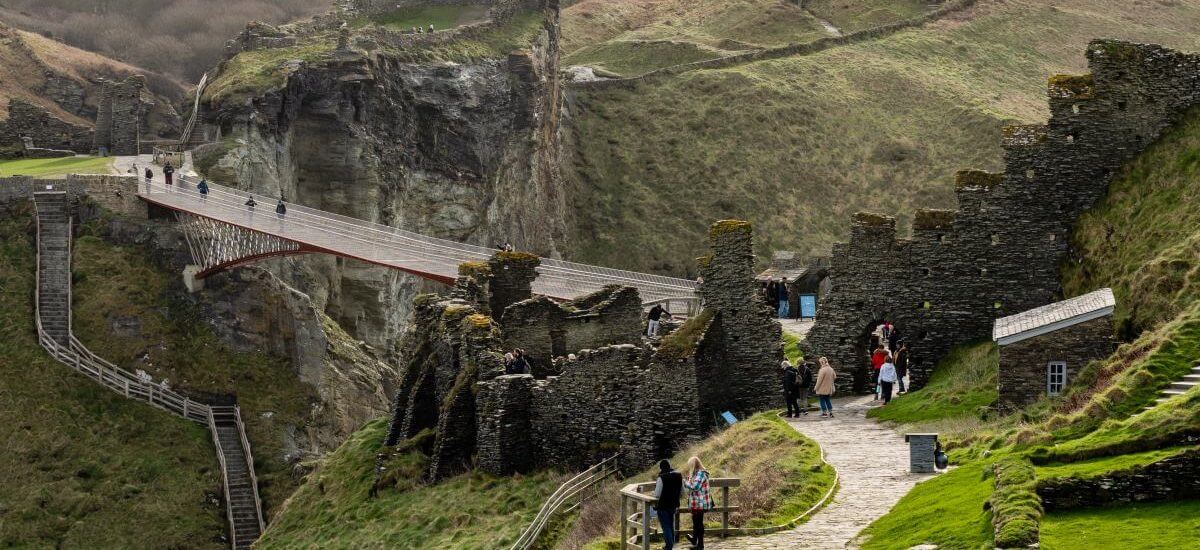 Cornwall, İngiltere'de bulunan ve Kral Arthur ile bağlantılı olduğuna inanılan Tintagel Kalesi kalıntılarının taş duvarlar, yeşil çimen, dolambaçlı yollar ve bulutlu bir gökyüzü altında dramatik bir uçurumun üzerindeki yaya köprüsünün yanında keşif yapan insanlarla birlikte doğal bir görünümü.