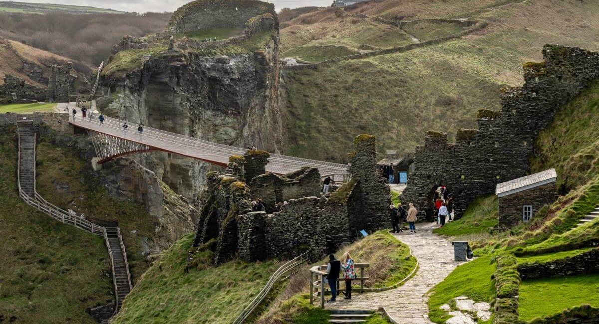 Cornwall, İngiltere'de bulunan ve Kral Arthur ile bağlantılı olduğuna inanılan Tintagel Kalesi kalıntılarının taş duvarlar, yeşil çimen, dolambaçlı yollar ve bulutlu bir gökyüzü altında dramatik bir uçurumun üzerindeki yaya köprüsünün yanında keşif yapan insanlarla birlikte doğal bir görünümü.