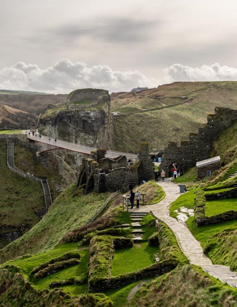 Cornwall, İngiltere'de bulunan ve Kral Arthur ile bağlantılı olduğuna inanılan Tintagel Kalesi kalıntılarının taş duvarlar, yeşil çimen, dolambaçlı yollar ve bulutlu bir gökyüzü altında dramatik bir uçurumun üzerindeki yaya köprüsünün yanında keşif yapan insanlarla birlikte doğal bir görünümü.
