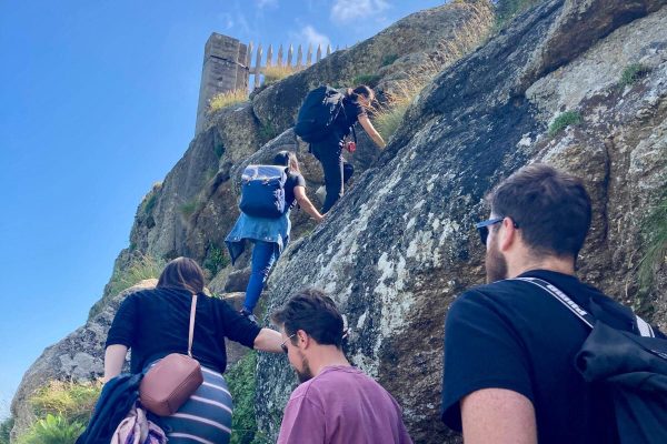 A group of people with backpacks climb stone steps along a rocky hillside under a blue sky, following the coast walk from Zennor to St Ives, with a fence visible at the top of the hill.