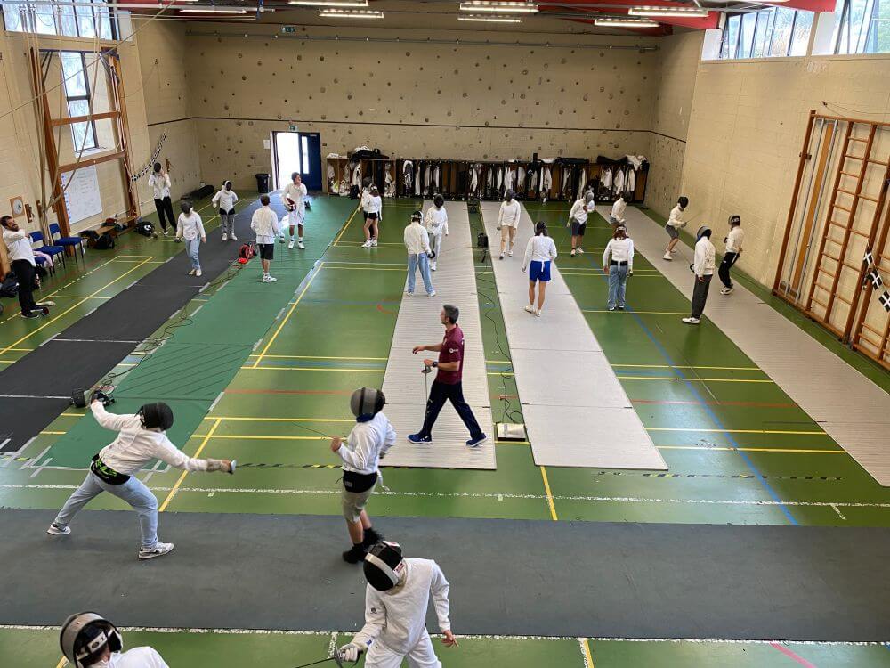 กลุ่มคนกำลังฝึกซ้อมฟันดาบในโรงยิมในร่มขนาดใหญ่ที่มีแถบฝึกฟันดาบหลายแถบและมีโค้ชเดินอยู่ระหว่างแถบฝึก คล้ายกับที่พบในการทัศนศึกษาอันน่าตื่นตาตื่นใจในคอร์นวอลล์ ผู้เข้าแข่งขันสวมชุดป้องกันและหน้ากากสีขาว โดยมีเสื้อแจ็คเก็ตแขวนอยู่ที่ผนังด้านหลัง