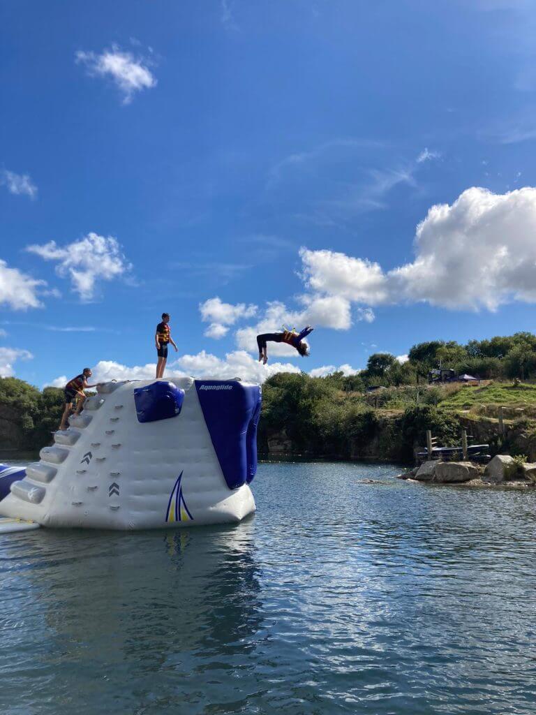 Drei Personen genießen einen Wassersporturlaub auf einer großen aufblasbaren Struktur in einem See; einer macht einen Rückwärtssalto in der Luft, während zwei andere zusehen, umgeben von strahlend blauem Himmel, vereinzelten Wolken und üppig grünen Bäumen.
