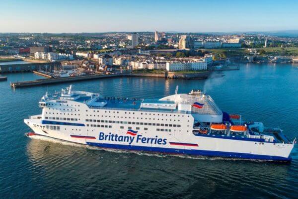 A large Brittany Ferries ship sails out of a harbor, heading towards the open sea, with a coastal city in the background under a clear blue sky—perfect for those travelling to Cornwall.
