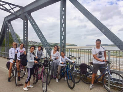 A group of eight people with bicycles pose and smile on a metal bridge overlooking a river or beach in Cornwall, enjoying social activities and excursions together under a cloudy sky. Some are standing, while others sit on their bikes.