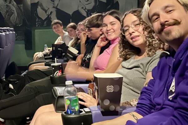 A group of people sit in a row of purple cinema seats, smiling at the camera during one of their social activities excursions in Cornwall. They have snacks and drinks while black and white portraits of classic film stars decorate the wall behind them.