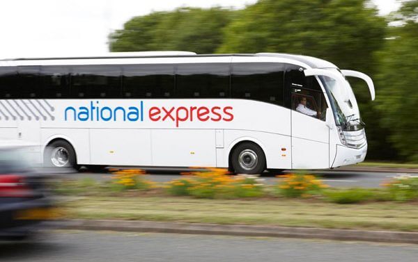 A white National Express coach bus, possibly travelling to Cornwall, drives on a road lined with grass and flowers, with trees and another blurred vehicle in the background.