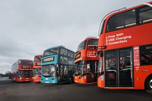 Five double-decker buses, mostly red and one blue, are parked side by side on a paved lot under a cloudy sky. The red bus in the foreground advertises free WiFi and USB charging—perfect for those travelling to Cornwall.