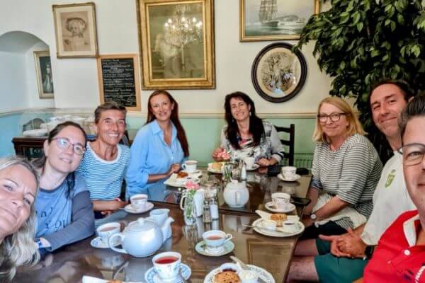A group of eight adults sit around a table in a cozy café in Cornwall, smiling at the camera. The table is set with teapots, cups of tea, and plates of scones and pastries—the perfect spot to relax after social activities or excursions.