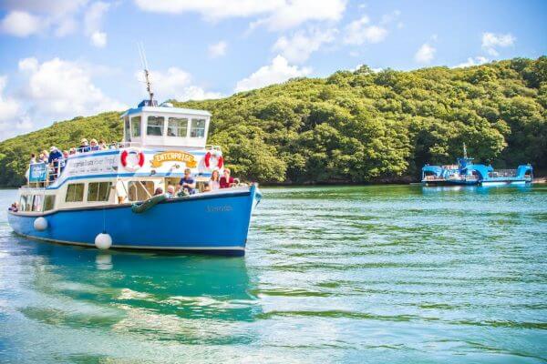 A blue and white passenger boat with people on board enjoys social activities while sailing on a greenish river in Cornwall, with another boat in the background and lush green trees lining the shore under a partly cloudy sky.