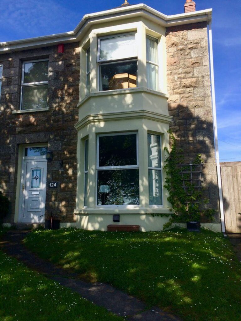 Stone house with large bay windows, white trim, and a white front door marked 124. Sunlight and tree shadows fall on the facade, highlighting the inviting accommodation options Cornwall has to offer. A green lawn sprinkled with small white flowers lies in front.