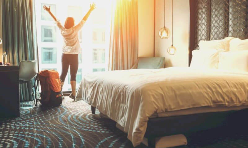 A person stands with arms raised toward a sunlit window in a modern hotel room, enjoying one of the many accommodation options Cornwall has to offer, with a backpack on the floor and a neatly made bed in the foreground.