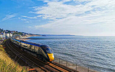A modern passenger train, travelling to Cornwall, glides along coastal railway tracks beside a calm sea, under a blue sky with scattered clouds and a distant shoreline in view.