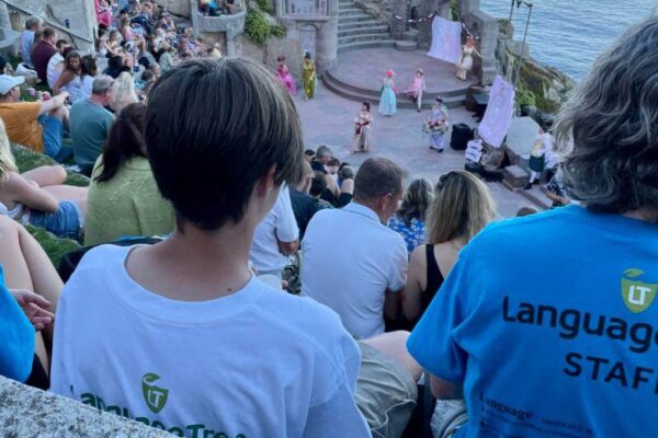 An audience enjoys an outdoor theater performance set on rocky cliffs by the sea in Cornwall. Some people wear Language Tree shirts, highlighting social activities and excursions as the stage, ocean, and cliffs create a stunning backdrop.