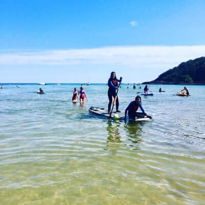 People enjoy social activities like paddleboarding and kayaking in calm, shallow sea water near a beach with a forested hill in the background under a clear blue sky, making for perfect excursions in Cornwall.