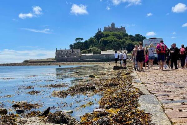 Tourists enjoy social activities and excursions as they walk along a stone causeway covered with seaweed at low tide, leading to a historic castle atop a green hill in Cornwall under a bright blue sky with scattered clouds.