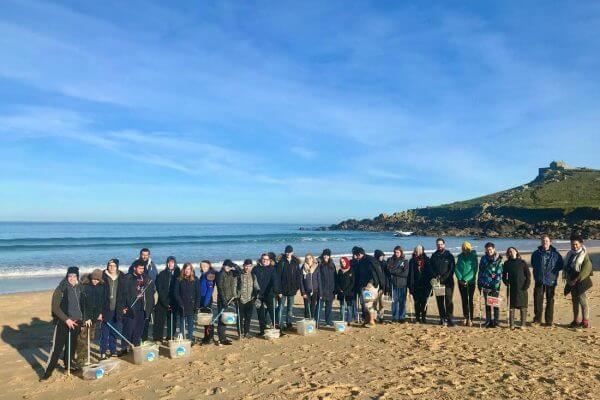 A large group of people stands on a sandy beach in Cornwall with the ocean behind them, holding buckets and litter pickers, participating in a beach cleanup excursion under a clear blue sky.