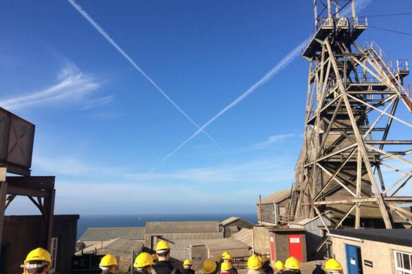 A group of people wearing yellow hard hats stand outside on an industrial site near the sea in Cornwall, with a metal tower and buildings nearby. Contrails form a large X overhead, marking their social activities excursion.