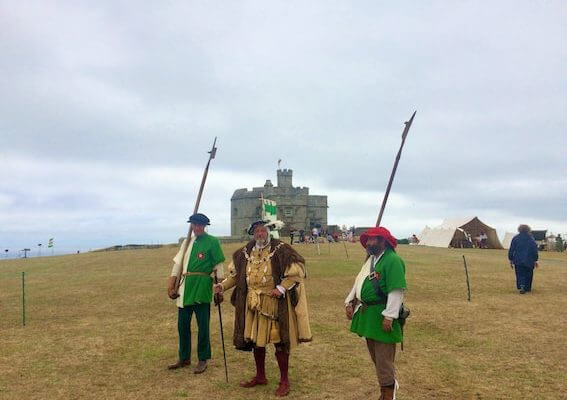 Three people in historical costumes stand on a grassy field holding spears during social activities excursions in Cornwall, with a stone castle and tents in the background under a cloudy sky.