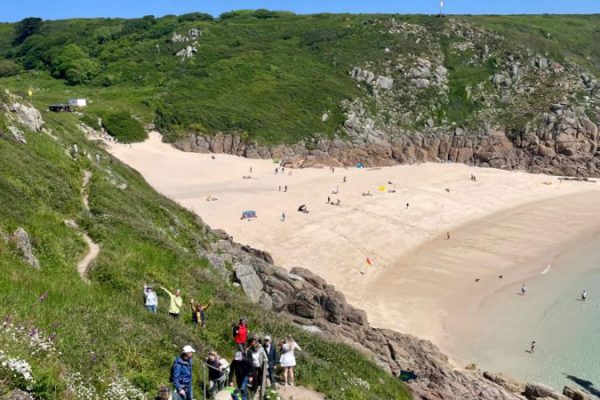 A group of people walk up a steep, rocky path lined with wildflowers overlooking a sandy beach and turquoise water, with grassy cliffs and a bright blue sky in the background.