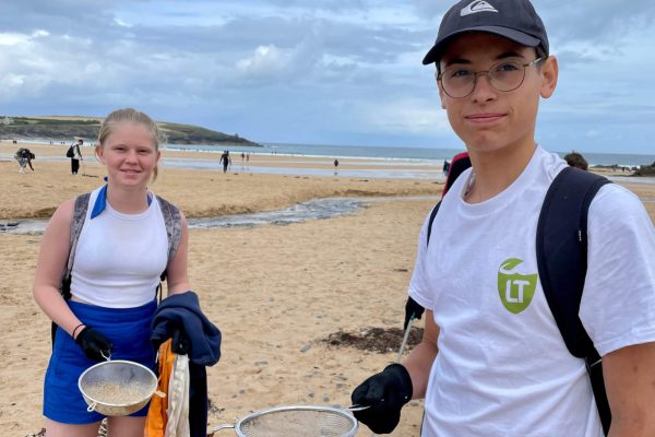 Two teenagers on a sandy beach hold metal sieves, possibly for beachcombing. The boy wears glasses, a black cap, and a white shirt with a green LT logo. The girl wears a white tank top and blue shorts. People and the ocean are in the background.