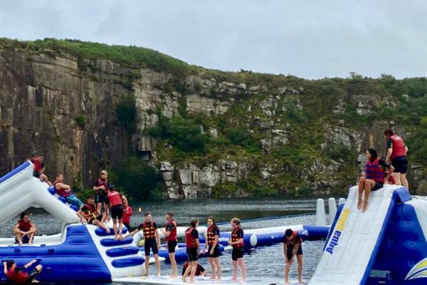 A group of people wearing life jackets enjoy social activities on a large inflatable obstacle course floating on a lake in Cornwall, with rocky cliffs and lush greenery as the backdrop. Some are climbing while others stand or swim nearby, making for fun excursions.