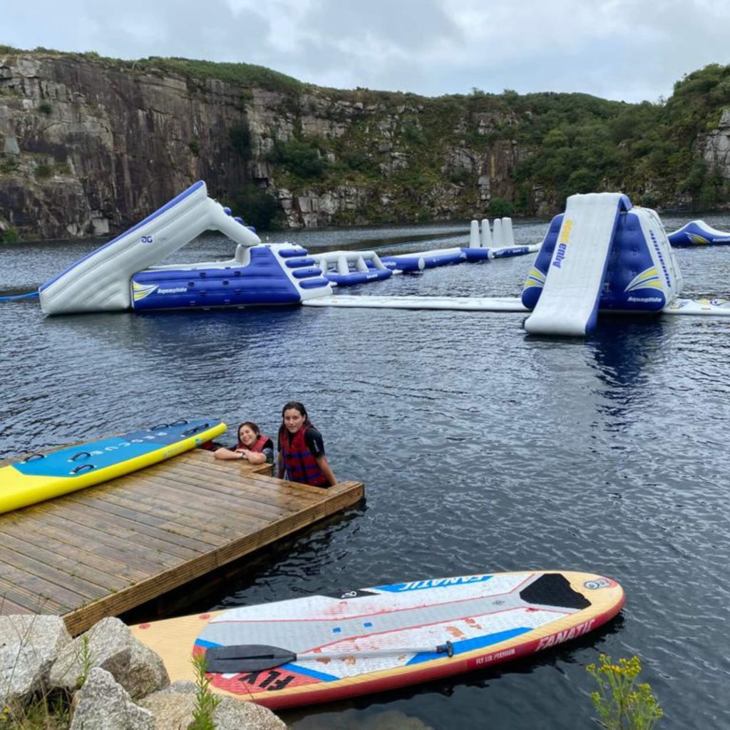 Two people in life jackets sit on a wooden dock beside paddleboards on a lake. Behind them, a large inflatable water obstacle course floats on the water, surrounded by rocky cliffs and greenery.