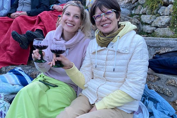 Two women sitting on stone steps outdoors, smiling and holding glasses of red wine. They are wrapped in warm clothes and blankets, with a group of people seated behind them. Mossy stone walls are visible in the background.