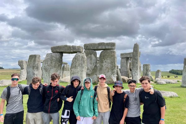 A group of eight teenagers pose together in front of Stonehenge under a cloudy sky, standing on green grass with the ancient stone monument in the background.
