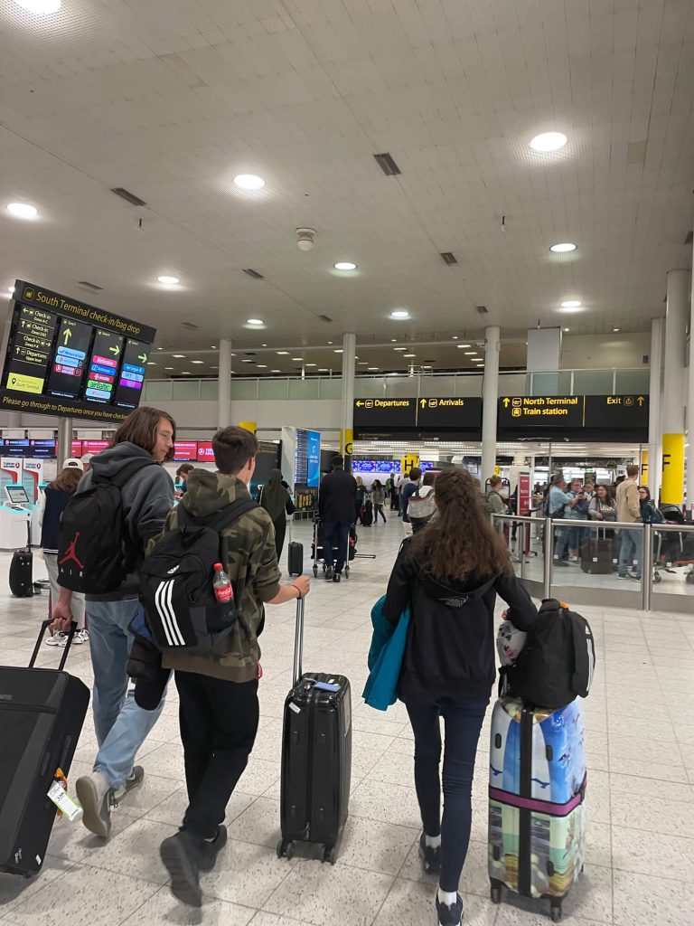 Travelers with suitcases walk through a busy airport terminal, with overhead signs for departures, arrivals, and train stations. Digital flight boards and other passengers are visible in the background.